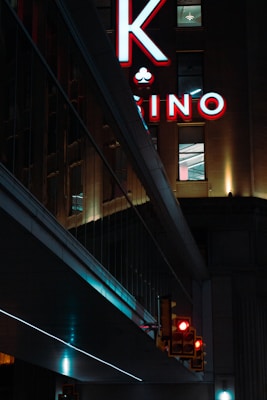 A night scene featuring a building with illuminated neon signage displaying 'CASINO'. The environment is urban with reflections on glass windows and glowing street traffic lights below. The atmosphere is dimly lit, with artificial lighting casting vibrant colors.