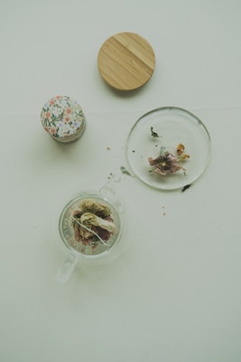 A transparent teapot containing dried flowers and leaves sits on a light-colored surface. Nearby are a glass lid with some dried flowers and leaves, a jar with a floral-patterned lid, and a wooden coaster.