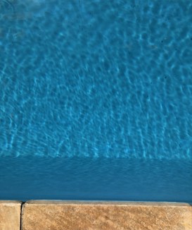 A section of a swimming pool with calm, rippling blue water. The water reflects varying shades of blue, creating a tranquil and refreshing appearance. At the bottom edge, there is a poolside deck composed of beige tiles.