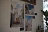 Three photographs of picturesque European streets are displayed on a light-colored wall. The photographs feature bright and sunny street scenes with white buildings, cobblestone paths, and decorative lamps. A leafy plant is placed nearby, adding greenery to the corner. The images on the wall appear to capture a charming and serene village environment.