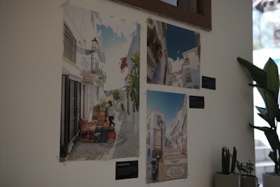 Three photographs of picturesque European streets are displayed on a light-colored wall. The photographs feature bright and sunny street scenes with white buildings, cobblestone paths, and decorative lamps. A leafy plant is placed nearby, adding greenery to the corner. The images on the wall appear to capture a charming and serene village environment.