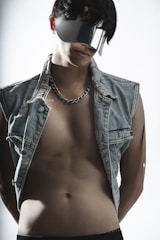 A person wearing futuristic, reflective sunglasses and a sleeveless denim jacket, exposing their torso. The lighting is soft, accentuating the textures of the clothing and the skin.