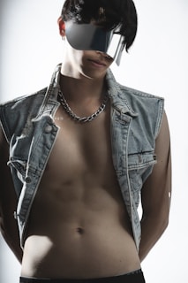 A person wearing futuristic, reflective sunglasses and a sleeveless denim jacket, exposing their torso. The lighting is soft, accentuating the textures of the clothing and the skin.