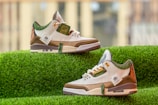 A pair of stylish sneakers with white, brown, and green accents is displayed on a vibrant green grass-like surface. The sneakers feature intricate designs on the heel and various labels and text on the side. The background is softly blurred, emphasizing the sneakers.