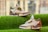 A pair of stylish sneakers with white, brown, and green accents is displayed on a vibrant green grass-like surface. The sneakers feature intricate designs on the heel and various labels and text on the side. The background is softly blurred, emphasizing the sneakers.