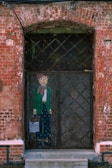 A painted figure of an anthropomorphic animal, dressed in stylish, vintage clothing, is depicted on a rustic metal door set within a red brick wall. The figure wears green jacket, scarf, and holds a bag with text. The bricks show signs of wear and age, adding a historical or urban decay aesthetic to the scene.