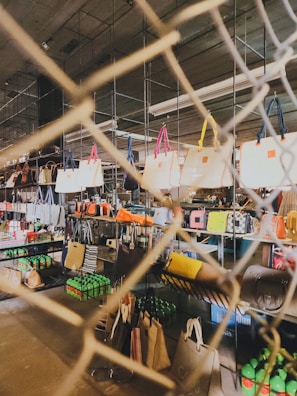 A variety of handbags are displayed on shelves behind a wire mesh. The bags have different colors and styles, hanging from metal racks in a well-lit indoor space. Below the bags, there are shelves stocked with green bottles and other miscellaneous items, suggesting a retail or market setting.