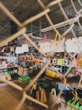 A variety of handbags are displayed on shelves behind a wire mesh. The bags have different colors and styles, hanging from metal racks in a well-lit indoor space. Below the bags, there are shelves stocked with green bottles and other miscellaneous items, suggesting a retail or market setting.