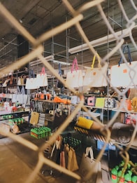 A variety of handbags are displayed on shelves behind a wire mesh. The bags have different colors and styles, hanging from metal racks in a well-lit indoor space. Below the bags, there are shelves stocked with green bottles and other miscellaneous items, suggesting a retail or market setting.