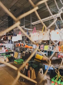 A variety of handbags are displayed on shelves behind a wire mesh. The bags have different colors and styles, hanging from metal racks in a well-lit indoor space. Below the bags, there are shelves stocked with green bottles and other miscellaneous items, suggesting a retail or market setting.