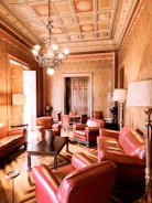 An opulently furnished room features richly upholstered red leather armchairs and sofas arranged around a dark wooden coffee table. The ceiling is intricately decorated with ornate moldings and a central chandelier. The walls are adorned with patterned wallpaper, and the flooring is a geometric parquet design. There is a wooden door at the far end, and several lamps provide additional lighting.