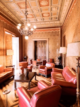 An opulently furnished room features richly upholstered red leather armchairs and sofas arranged around a dark wooden coffee table. The ceiling is intricately decorated with ornate moldings and a central chandelier. The walls are adorned with patterned wallpaper, and the flooring is a geometric parquet design. There is a wooden door at the far end, and several lamps provide additional lighting.