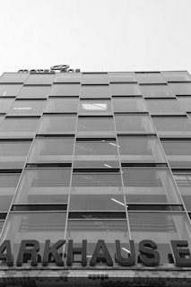 A multi-story glass building with a modern design, featuring a grid of large windows. A partially visible sign at the top reads 'MOTEL ONE', and at the lower portion, a large sign displays 'PARKHAUS'. The structure appears to be utilitarian with straight lines and reflective surfaces.