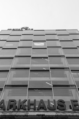 A multi-story glass building with a modern design, featuring a grid of large windows. A partially visible sign at the top reads 'MOTEL ONE', and at the lower portion, a large sign displays 'PARKHAUS'. The structure appears to be utilitarian with straight lines and reflective surfaces.