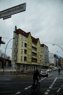 An urban street scene featuring a multi-story residential building with a mixture of gray and yellow facades. The building has several balconies and windows. In the foreground, there are pedestrians and cyclists navigating a crosswalk, with cars driving on the wet road. Overhead electric wires suggest the presence of a tram system, and there is a street sign with the text 'arnweberstraße'. A cloudy sky is visible above.
