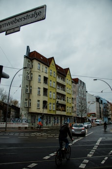 An urban street scene featuring a multi-story residential building with a mixture of gray and yellow facades. The building has several balconies and windows. In the foreground, there are pedestrians and cyclists navigating a crosswalk, with cars driving on the wet road. Overhead electric wires suggest the presence of a tram system, and there is a street sign with the text 'arnweberstraße'. A cloudy sky is visible above.