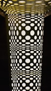 A geometric pattern composed of numerous circular holes in varying sizes, creating an optical illusion effect. The structure appears illuminated from behind, casting light through the holes and creating a contrast between light and shadows.
