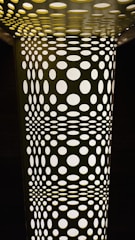 A geometric pattern composed of numerous circular holes in varying sizes, creating an optical illusion effect. The structure appears illuminated from behind, casting light through the holes and creating a contrast between light and shadows.