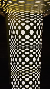 A geometric pattern composed of numerous circular holes in varying sizes, creating an optical illusion effect. The structure appears illuminated from behind, casting light through the holes and creating a contrast between light and shadows.