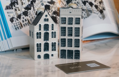 Two small, ceramic model houses are displayed on a smooth surface. They are detailed with windows and doors, resembling traditional European architecture. A credit card is placed in front of them. In the background, there is an open book showing similar house models arranged in a pattern.