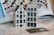 Two small, ceramic model houses are displayed on a smooth surface. They are detailed with windows and doors, resembling traditional European architecture. A credit card is placed in front of them. In the background, there is an open book showing similar house models arranged in a pattern.