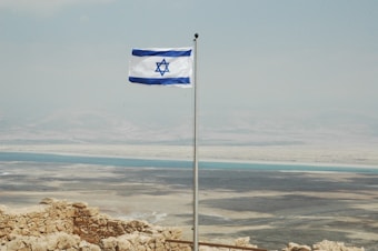 A flag featuring a blue Star of David between two horizontal blue stripes is waving on a flagpole. In the background, a vast desert landscape stretches out with the Dead Sea visible under a hazy sky.