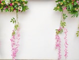 Colorful floral arrangements hang against a white background, featuring a variety of leaves and flowers in shades of green, pink, purple, and white. The arrangement includes cascading pink flowers that hang gracefully alongside vibrant green leaves.