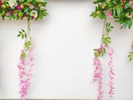 Colorful floral arrangements hang against a white background, featuring a variety of leaves and flowers in shades of green, pink, purple, and white. The arrangement includes cascading pink flowers that hang gracefully alongside vibrant green leaves.
