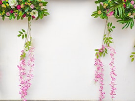 Colorful floral arrangements hang against a white background, featuring a variety of leaves and flowers in shades of green, pink, purple, and white. The arrangement includes cascading pink flowers that hang gracefully alongside vibrant green leaves.