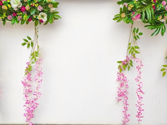 Colorful floral arrangements hang against a white background, featuring a variety of leaves and flowers in shades of green, pink, purple, and white. The arrangement includes cascading pink flowers that hang gracefully alongside vibrant green leaves.