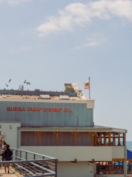 A building with signage for 'Bubba Gump Shrimp Co.' located on a pier. The structure has a flag pole with a California state flag and is populated by several birds perched on the roof. A railing leads up to the building, and people are walking along the walkway.