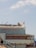 A building with signage for 'Bubba Gump Shrimp Co.' located on a pier. The structure has a flag pole with a California state flag and is populated by several birds perched on the roof. A railing leads up to the building, and people are walking along the walkway.