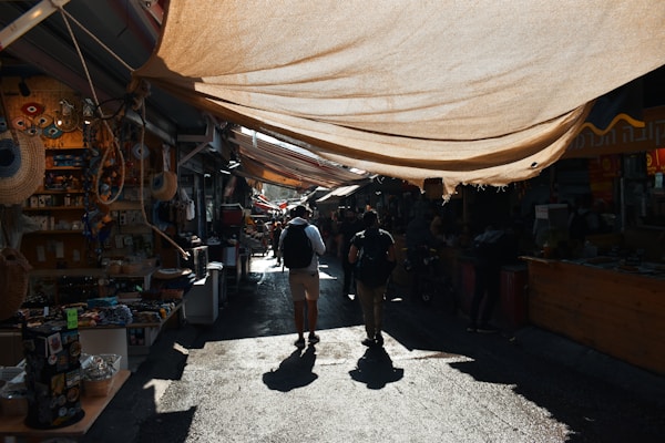 A lively marketplace with stalls along both sides of a narrow path, covered by large fabric canopies that cast shadows. People are walking through the market, browsing various goods such as souvenirs, fabrics, and local crafts displayed at the stalls.