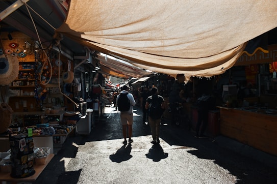 A lively marketplace with stalls along both sides of a narrow path, covered by large fabric canopies that cast shadows. People are walking through the market, browsing various goods such as souvenirs, fabrics, and local crafts displayed at the stalls.