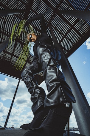 A person wearing futuristic, oversized black leather attire poses dramatically under a structure with a perforated roof. Their outfit includes tall boots and pants, creating a bold and avant-garde look. They wear dark sunglasses and hold what appears to be long, green plant tendrils, adding an artistic flair. The sky is partially visible, showing scattered clouds.