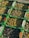 Several green crates are filled with olives of varying colors, including shades of green, purple, and black. The crates are stacked, displaying a rich, abundant harvest. Some olives are mixed with leaves or stems, suggesting a fresh, recently picked collection.