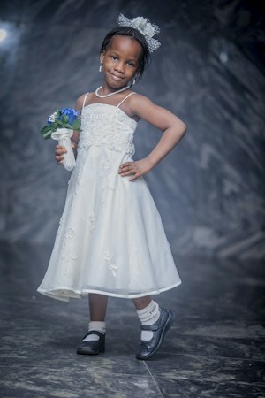A young girl stands confidently, wearing a white dress with floral patterns. She holds a small bouquet of blue and white flowers and has a delicate headband with flowers. The background is a soft, dark marbled texture, enhancing the elegant feel.