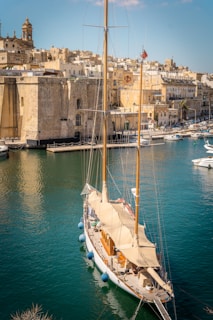 A large, elegant sailboat is docked in a calm harbor bordered by historic stone buildings. The structures have a warm beige tone, reflecting the sunlight, with a tower visible on the left. The vibrant blue-green water creates a striking contrast with the light-colored boat.