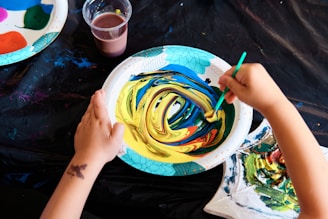 A child's hands are seen engaging in a painting activity. One hand holds a brush with a green handle while swirling a mixture of yellow, blue, and red paint on a paper plate. Nearby, another plate with a variety of paint colors and a cup with brownish liquid are visible. The background includes a black tablecloth with paint smudges.