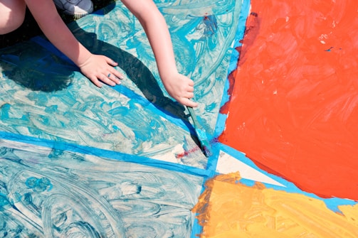 A child's hands are painting a large surface with various sections filled with bold colors, including blue, red, and orange. The painting appears abstract with swirling patterns and sections divided by blue tape.