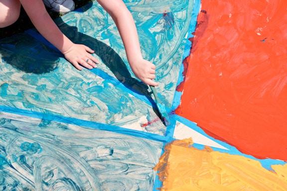 A child's hands are painting a large surface with various sections filled with bold colors, including blue, red, and orange. The painting appears abstract with swirling patterns and sections divided by blue tape.