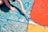 A child's hands are painting a large surface with various sections filled with bold colors, including blue, red, and orange. The painting appears abstract with swirling patterns and sections divided by blue tape.