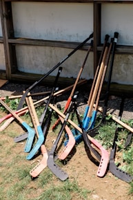 A pile of colorful street hockey sticks rests against a wooden structure. The sticks have various colors, including blue, red, and black blades. The scene includes patches of grass and dirt around the base of the sticks.