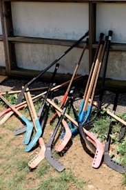 A pile of colorful street hockey sticks rests against a wooden structure. The sticks have various colors, including blue, red, and black blades. The scene includes patches of grass and dirt around the base of the sticks.