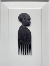 A carved wooden comb featuring the side profile of a person's head and upper body. The intricate design includes detailed facial features and decorative patterns on the torso, likely representing traditional or cultural art.
