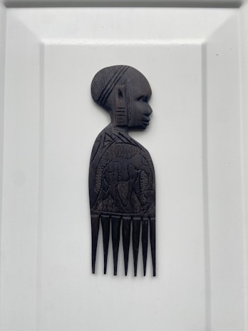 A carved wooden comb featuring the side profile of a person's head and upper body. The intricate design includes detailed facial features and decorative patterns on the torso, likely representing traditional or cultural art.
