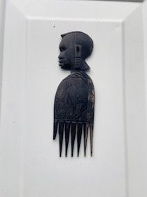 A carved wooden comb featuring an intricately designed profile of a person's head at the top. The comb showcases detailed features in the profile with hair, facial characteristics, and patterns on the handle.