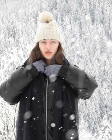 A person stands in a snowy environment, wearing a thick black coat, grey gloves, and a cream-colored knitted beanie with a pompom. The backdrop features snow-covered trees, and snow is falling, creating a serene winter scene.