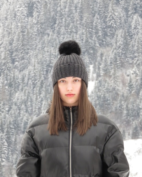 A person wearing a dark winter jacket and a knitted beanie with a pom-pom stands in front of a snowy forest landscape. The background consists of densely packed snow-covered trees, creating a serene winter setting.