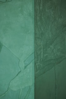 A textured wall with varying shades of green, showing both smoother and more rugged surfaces. The left side of the wall has lighter green tones, while the right side features a darker, deeper green with visible textures and patterns.